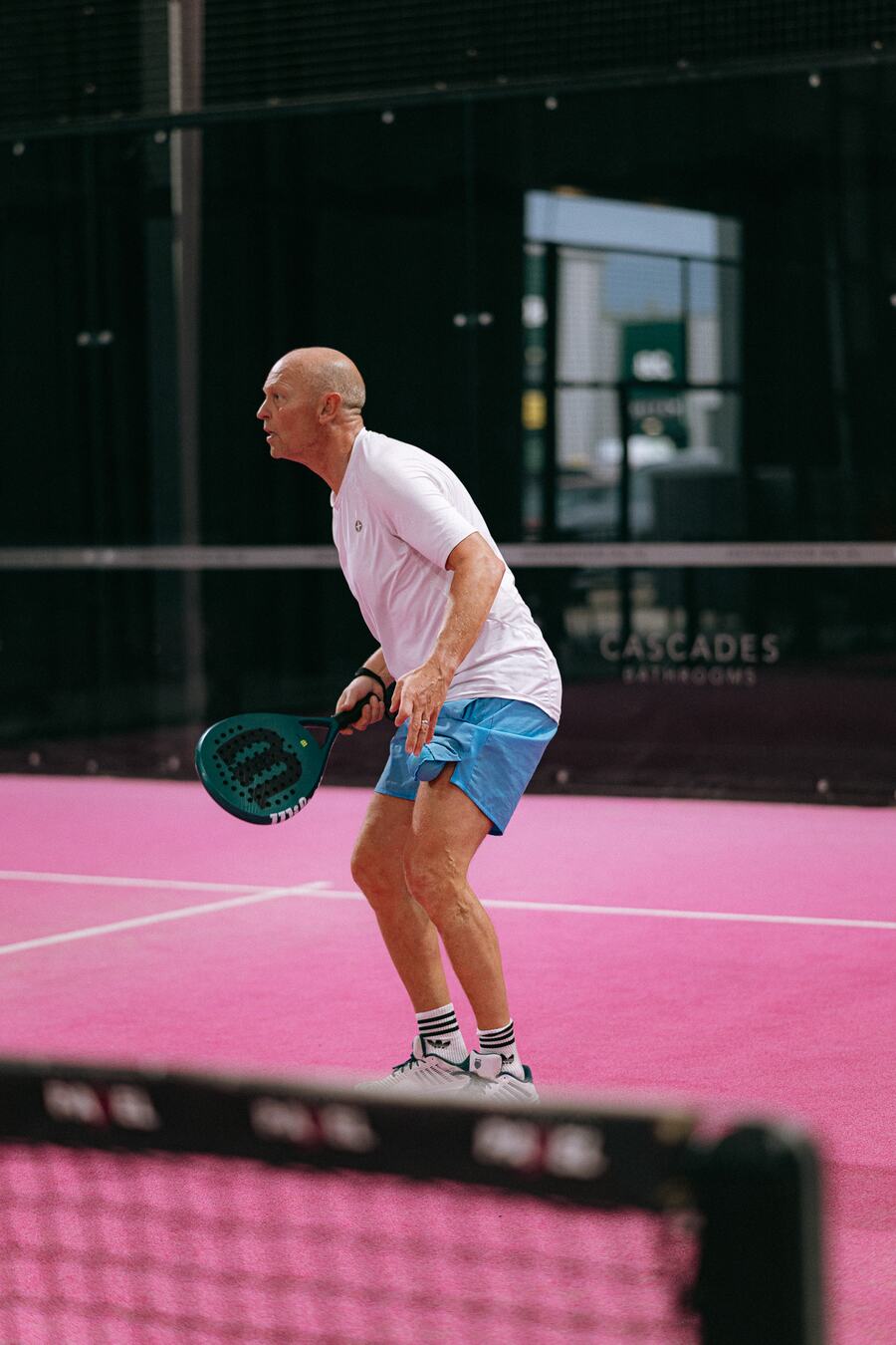 A player in white crouched and ready to return, seen from the side.