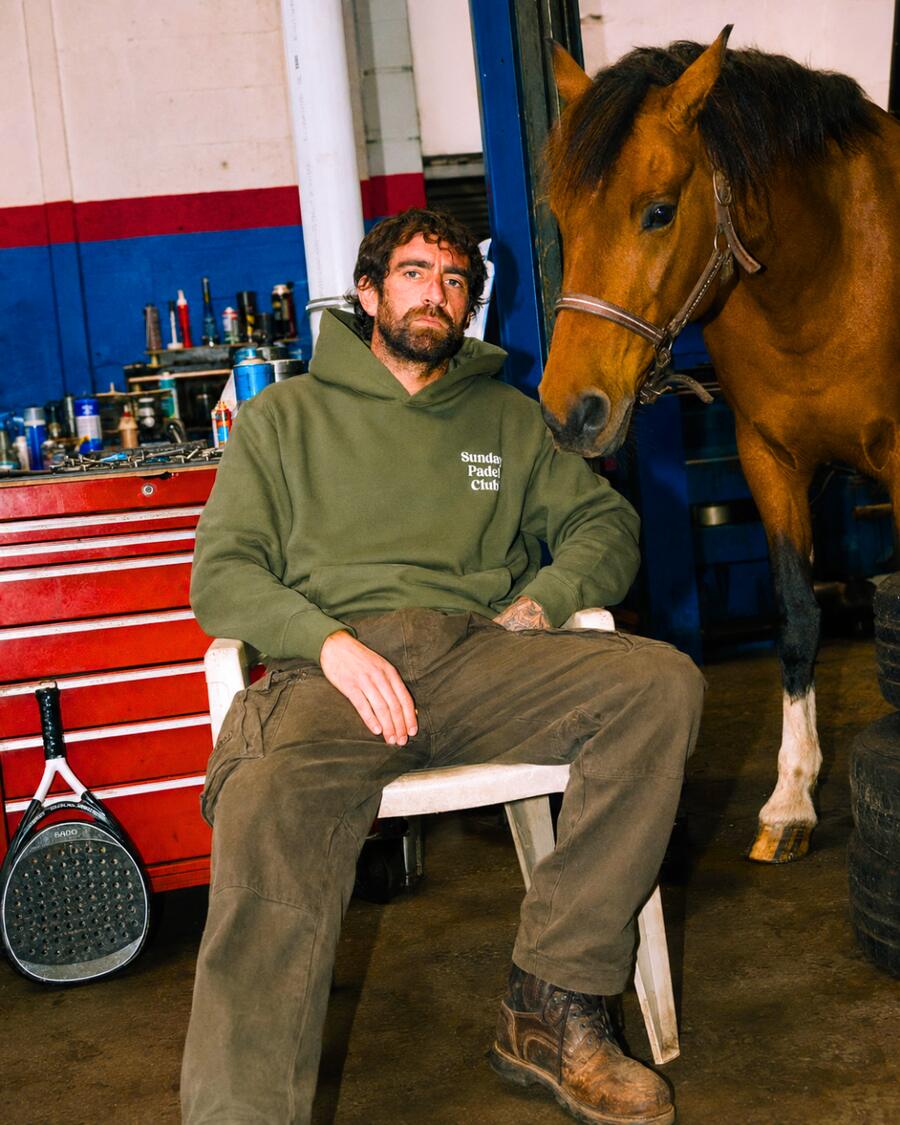 A bearded man in an olive Sunday Padel Club hoodie sitting in a garage next to a horse.