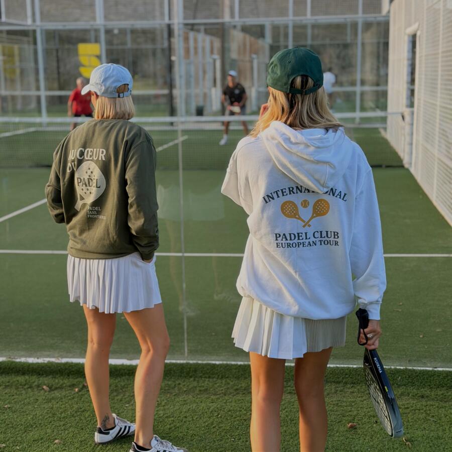 Two players in PSF hoodies and pleated skirts standing on a padel court, seen from behind.
