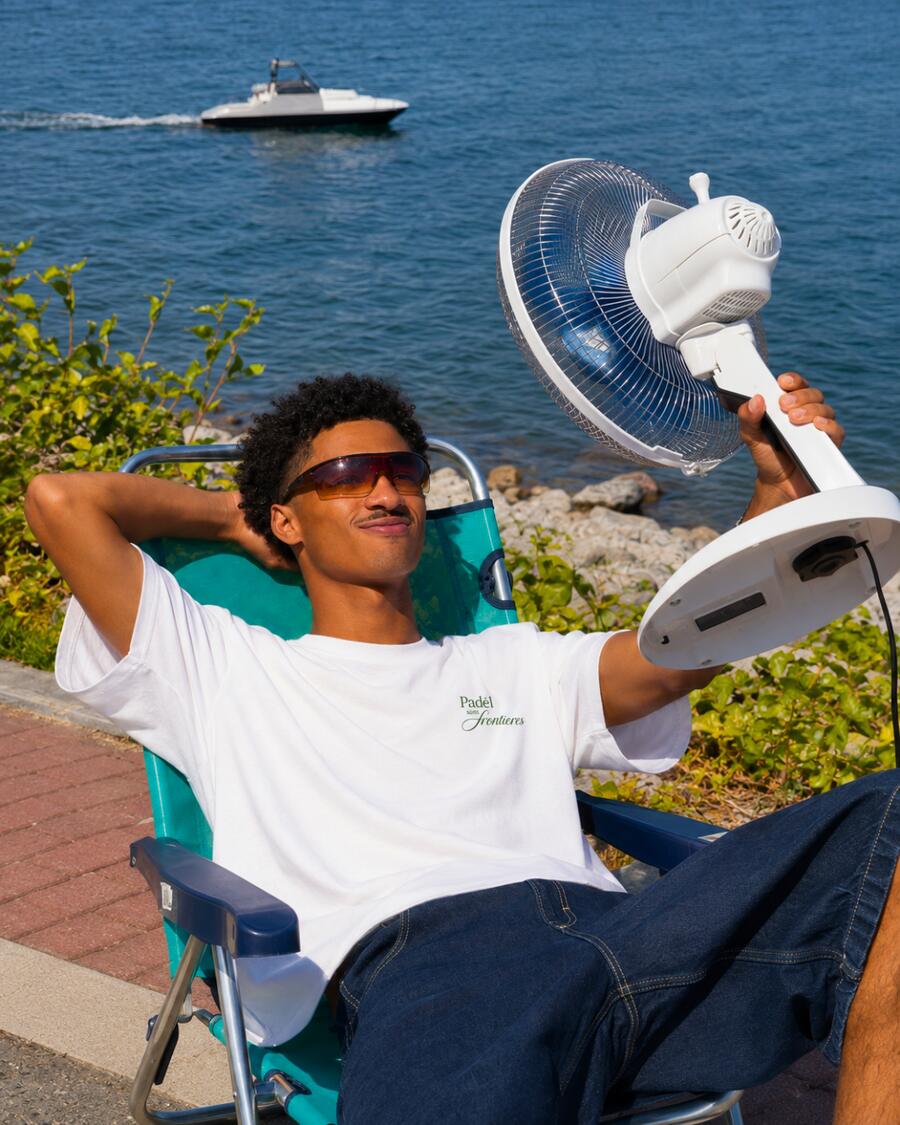 A man in a white PSF T-shirt and sunglasses relaxing in a deck chair by the sea, holding a fan.