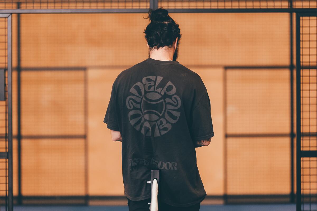 The back of a Vida Del Padel ambassador T-shirt, worn by a player facing an orange court wall.