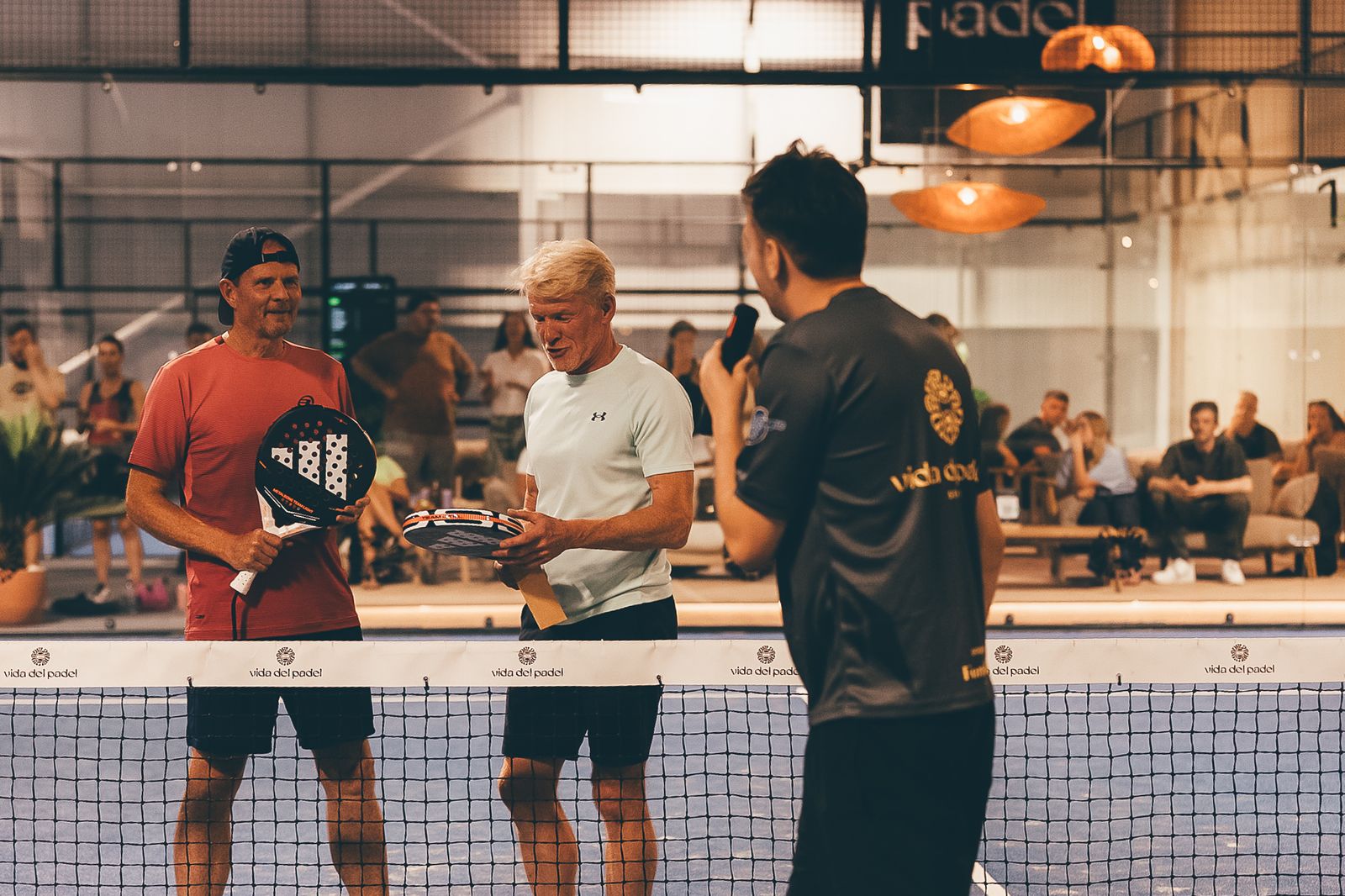 Two spectators at the net, holding rackets, with a host holding a microphone, after winning a challenge at a Vida Del Padel final.