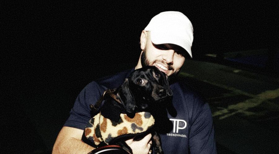 A smiling player in a white cap holding a small dog in a spotted coat, with a padel racket.