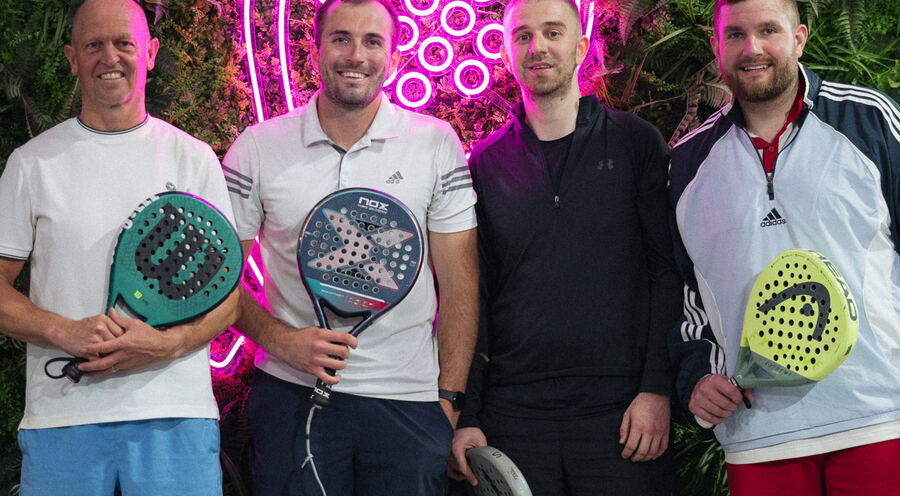 Four players holding padel rackets in front of a green wall with a pink neon racket sign.