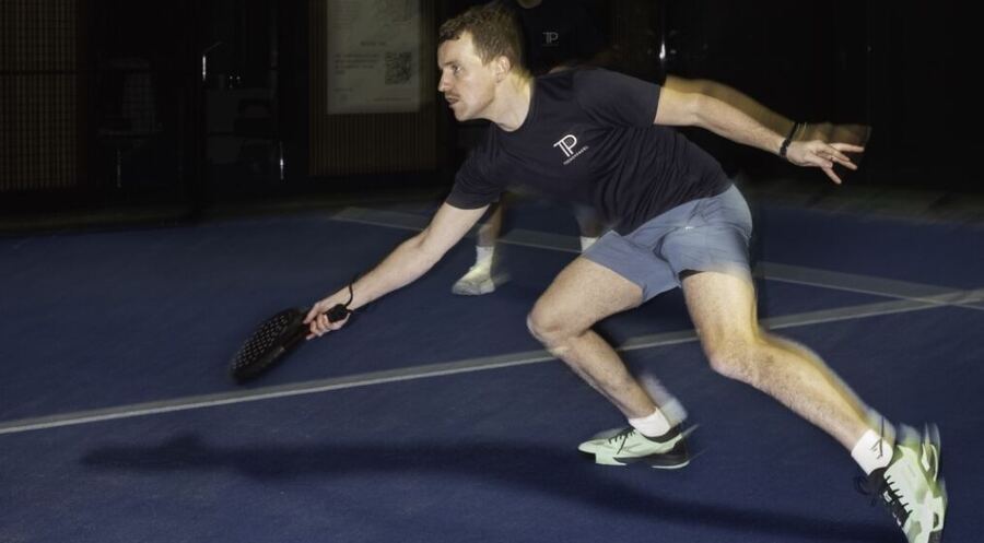 A player lunging low for a shot on a blue indoor padel court.