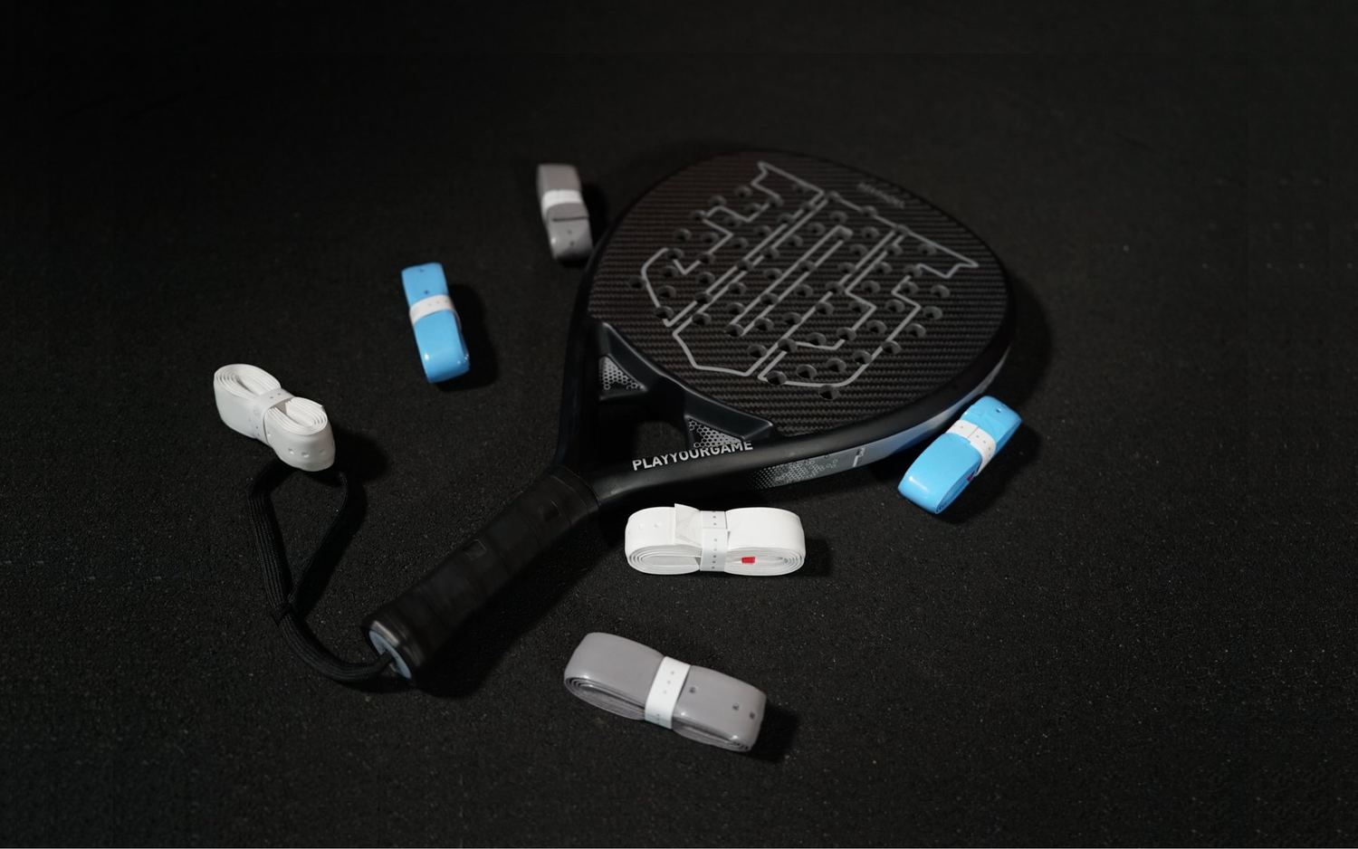 A black padel racket with its wrist strap, with pale blue, white and grey grips scattered around it on a dark background.