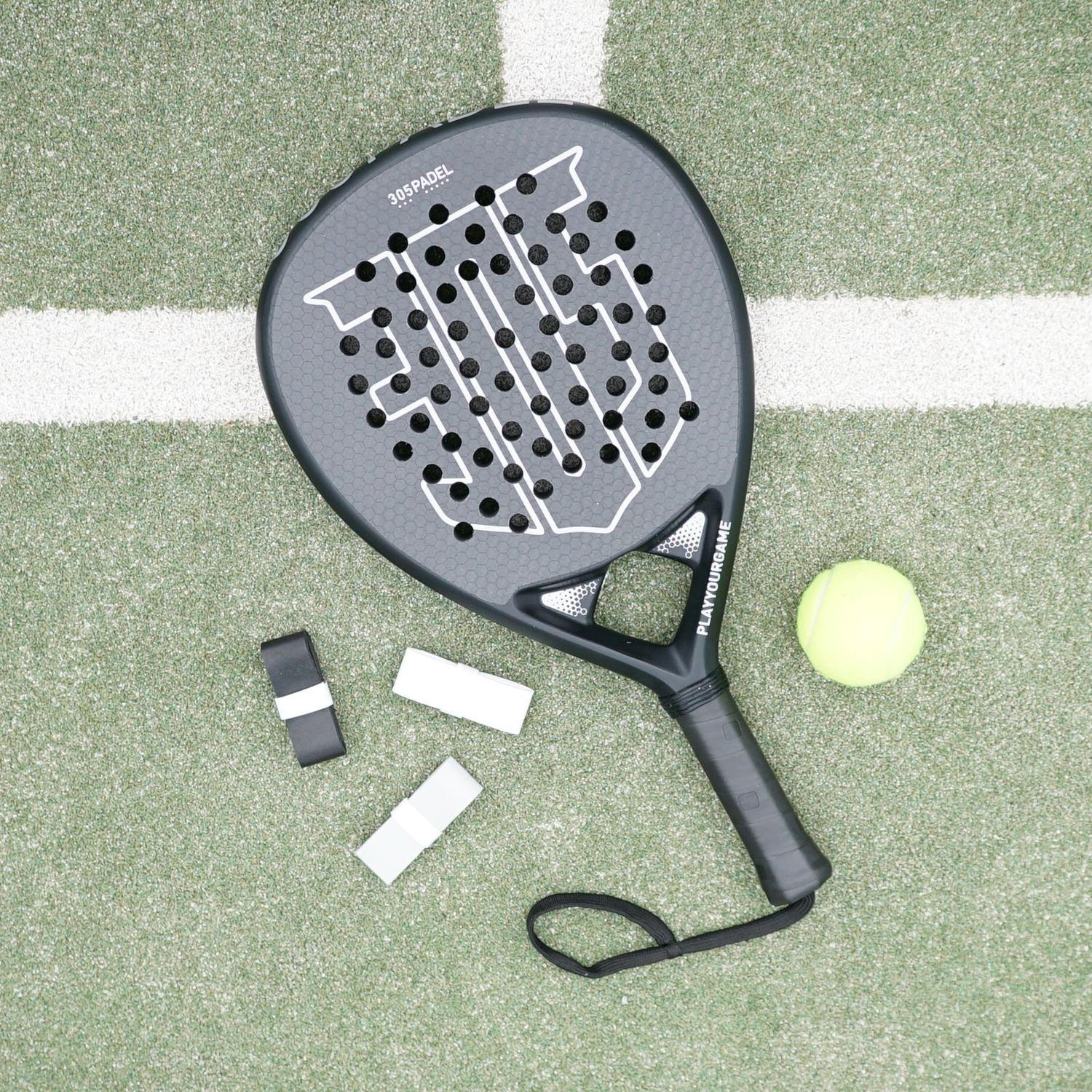 A grey padel racket with two grips and a yellow ball resting on a green court beside a white line.