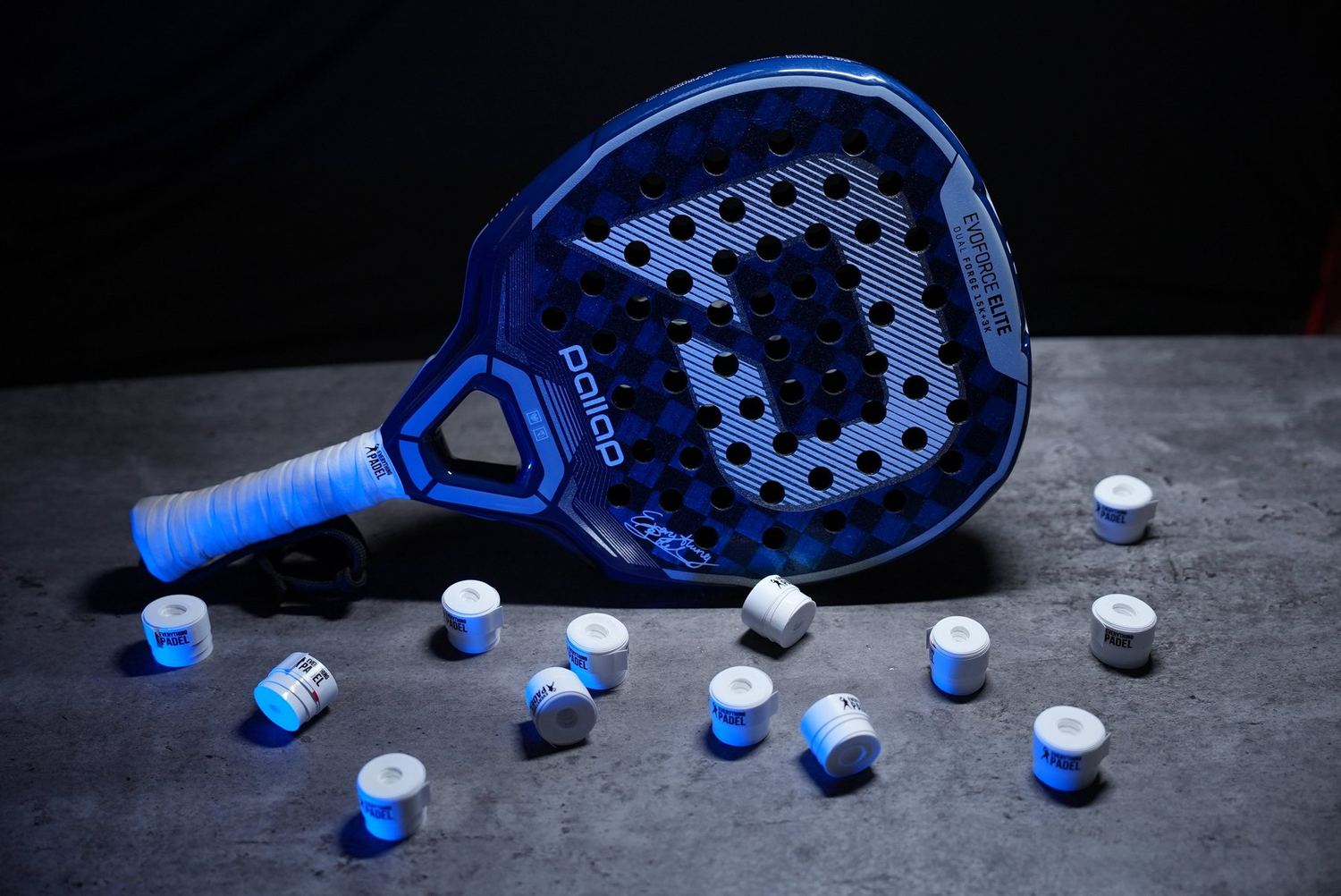 A blue and white padel racket lying on a dark surface with a scatter of overgrips around it.