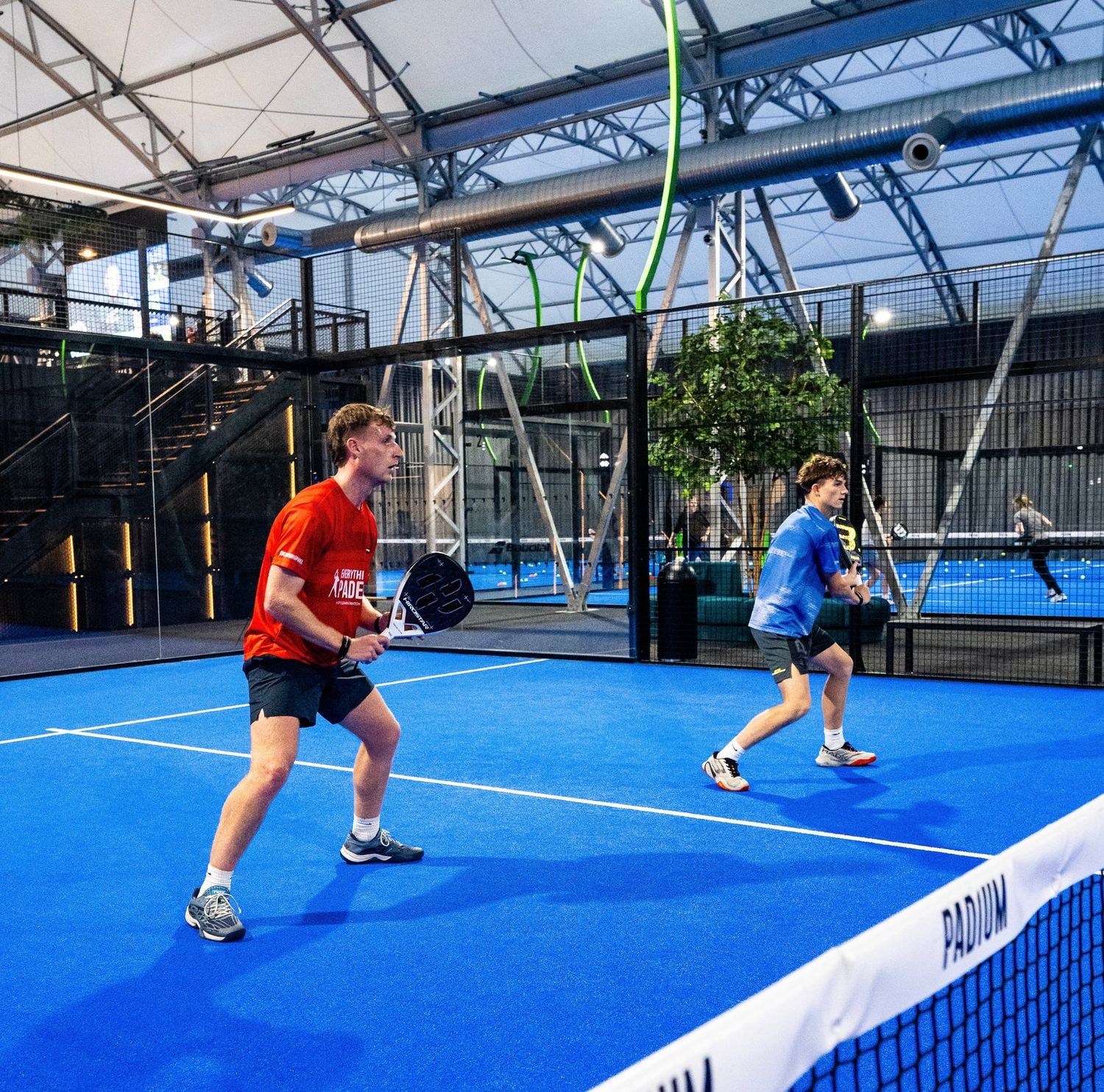 Two players in a rally on a blue indoor padel court under a high roof.