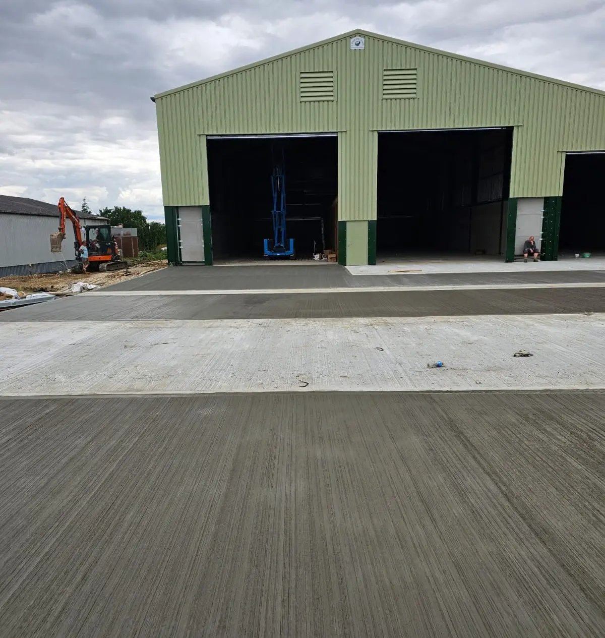 A green agricultural building with open doors and a brushed concrete apron in front.