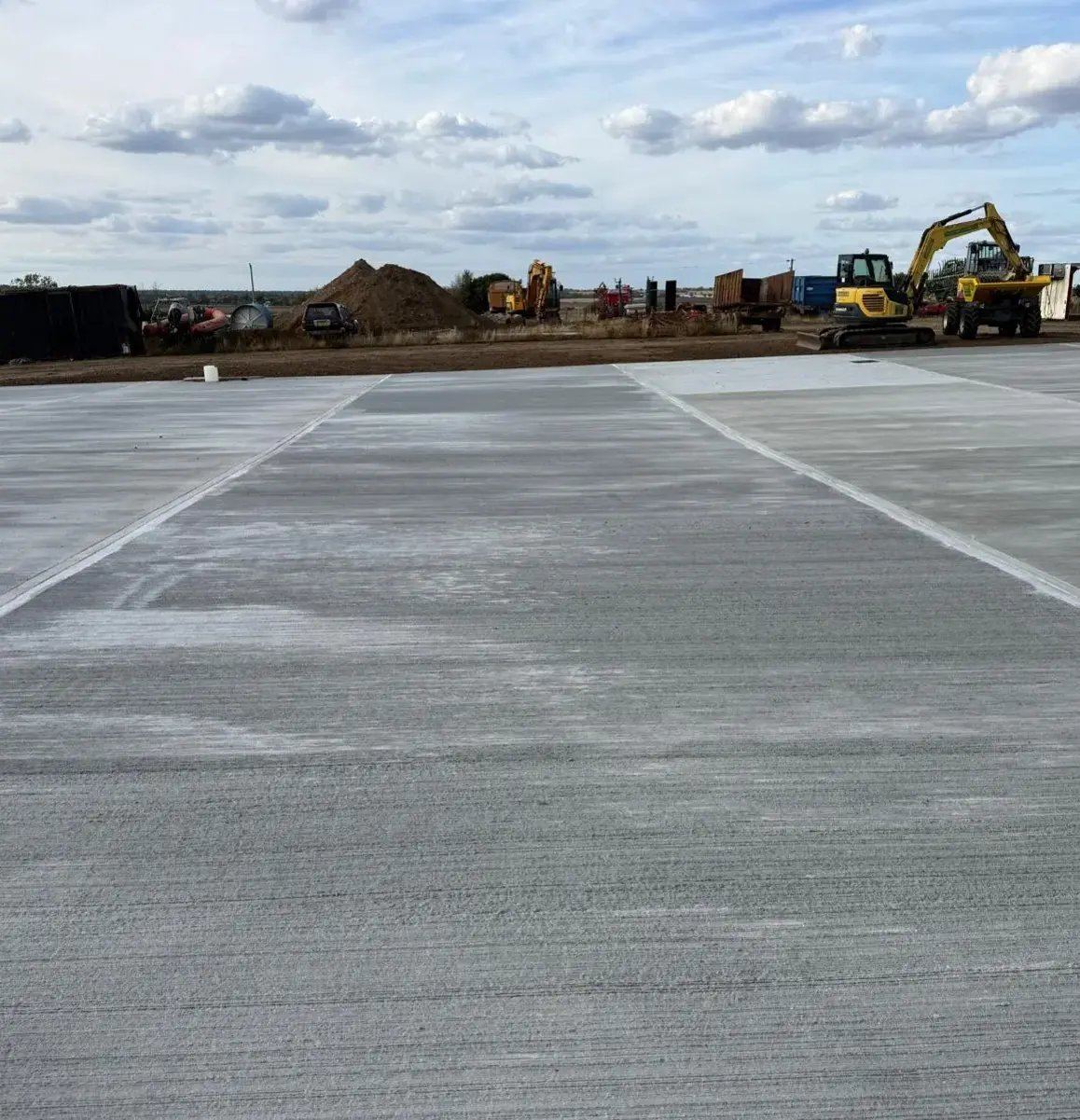 A wide concrete slab with a textured finish and digging machines working in the distance under a cloudy sky.