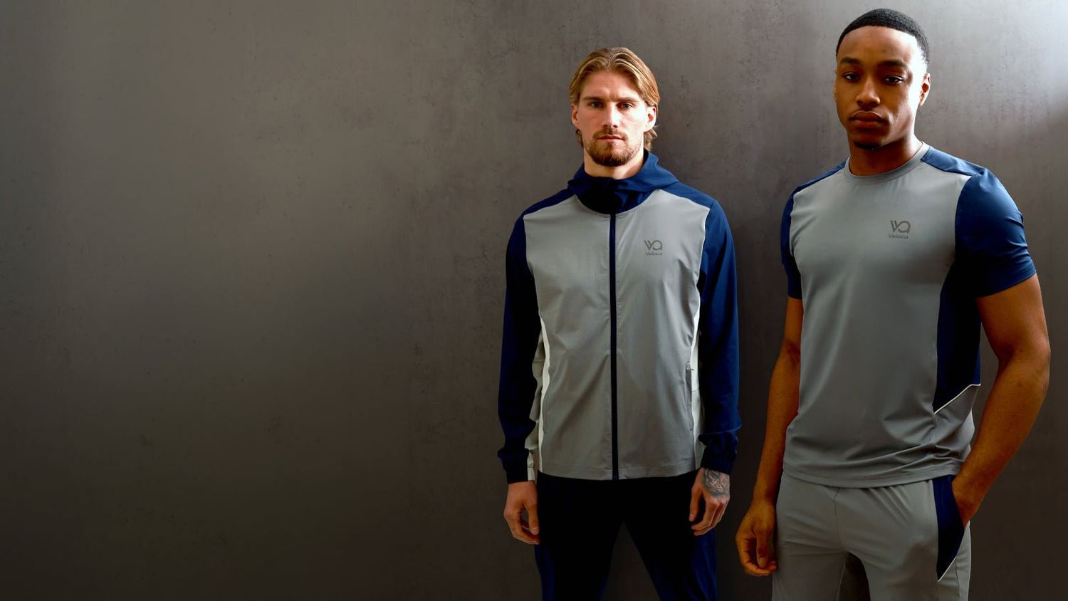 Two men in grey and navy Evolve jacket and t-shirt standing against a dark studio wall.