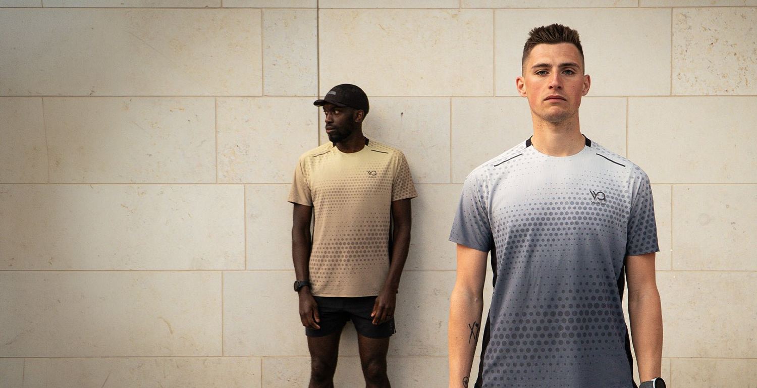 Two men in Performance t-shirts, one sand coloured and one grey fading to white, standing against a pale stone wall.