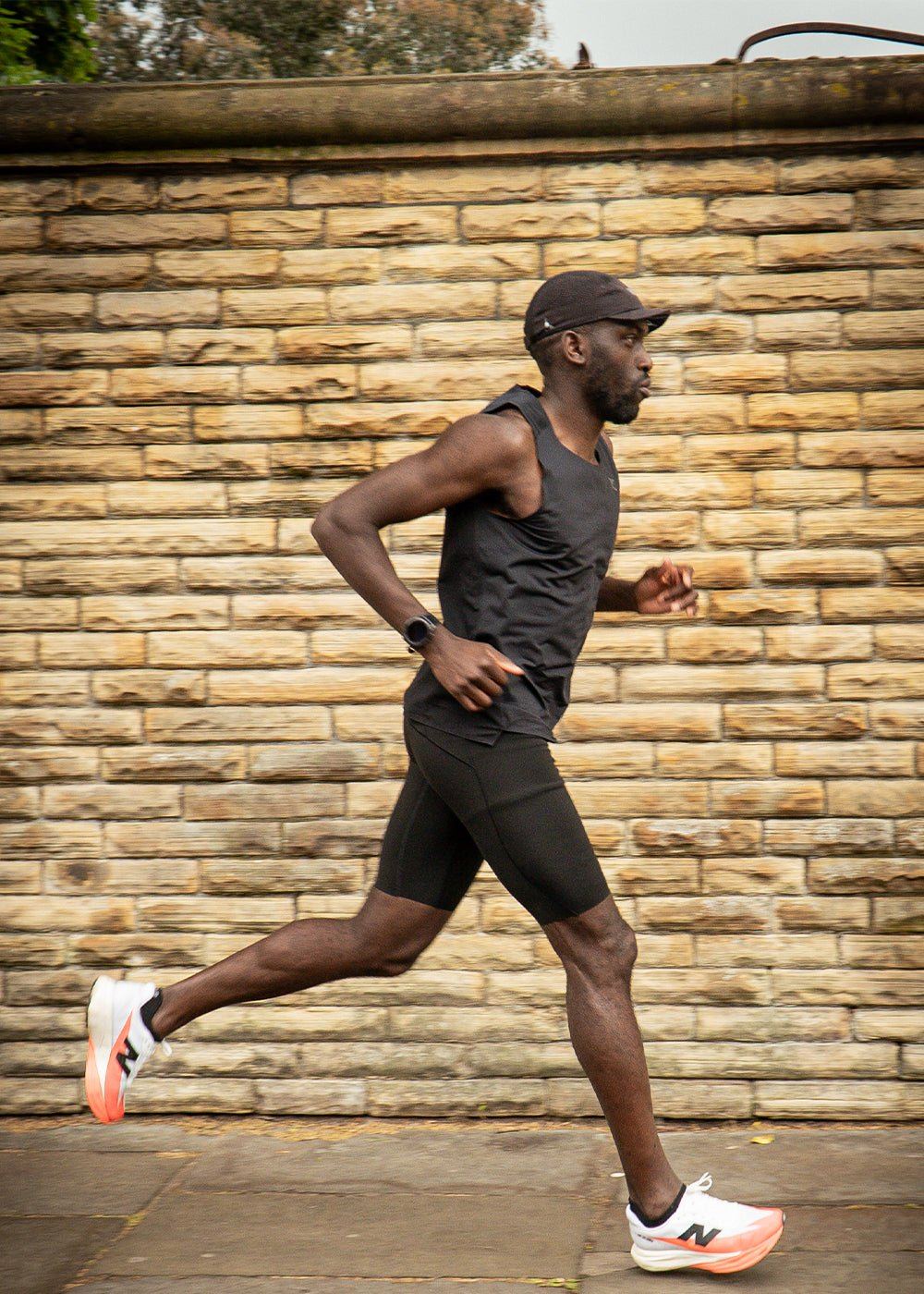 A runner in a black vest and shorts mid-stride in front of a sandstone wall.
