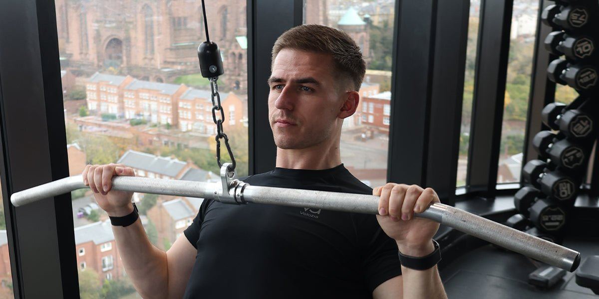 A man in a black t-shirt pulling a cable bar in a gym, with a view over a city behind him.