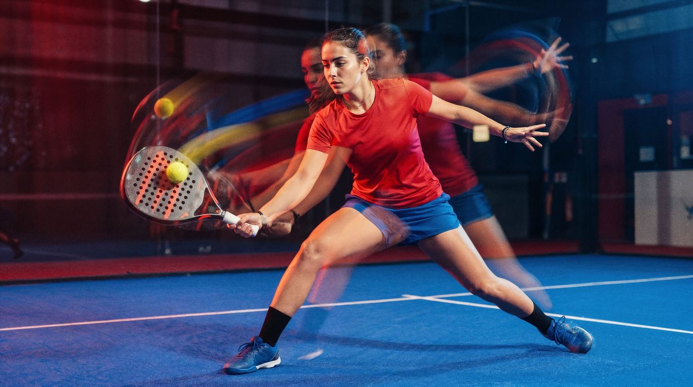 A player in a red top lunging to return a ball on a blue indoor padel court, shown with motion blur