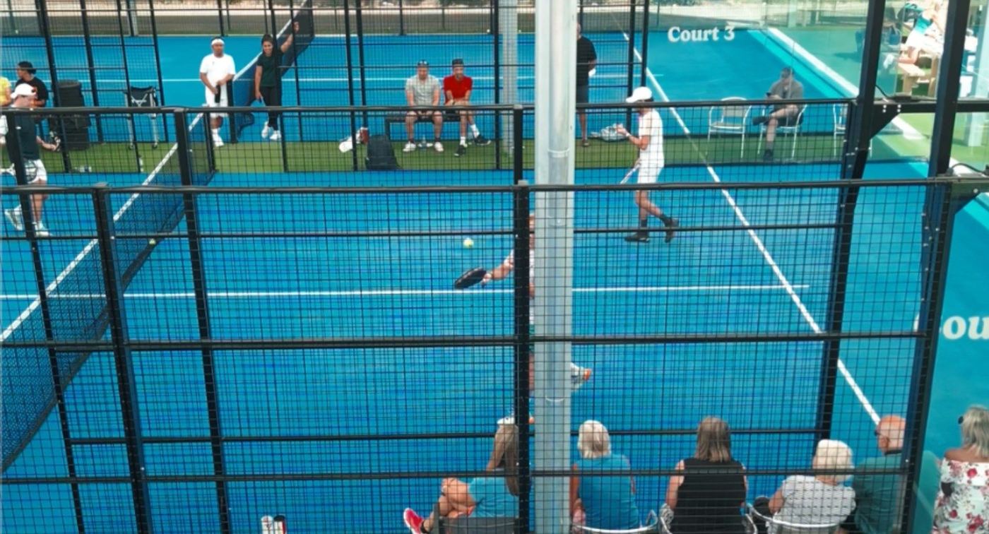 Blue padel courts seen through the fencing, with people watching and playing
