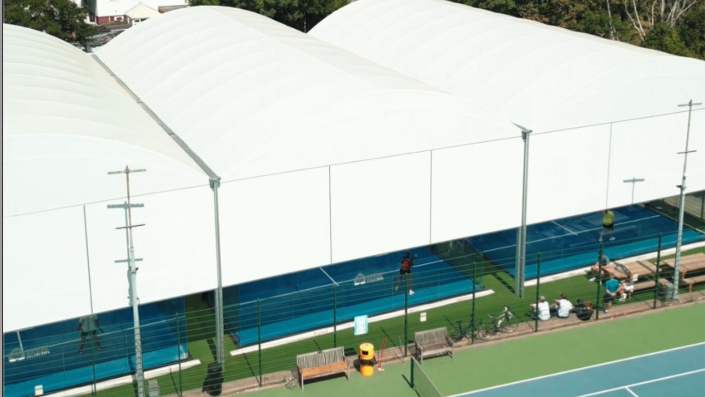 A large white canopy roof over padel courts, seen from above, with green space beside it