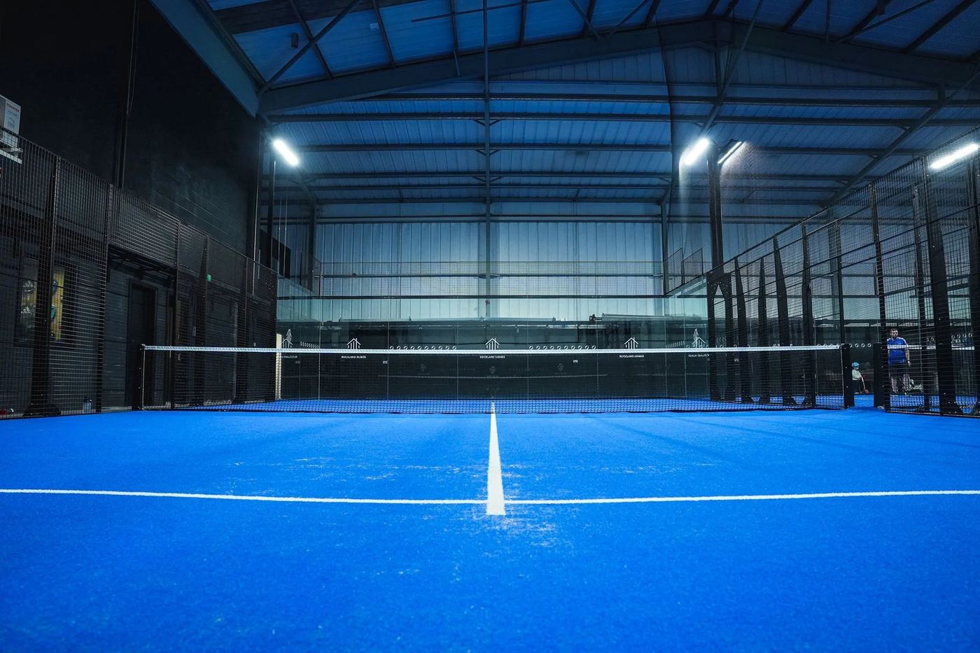 A blue indoor padel court viewed along the centre line, with high steel roof and floodlights