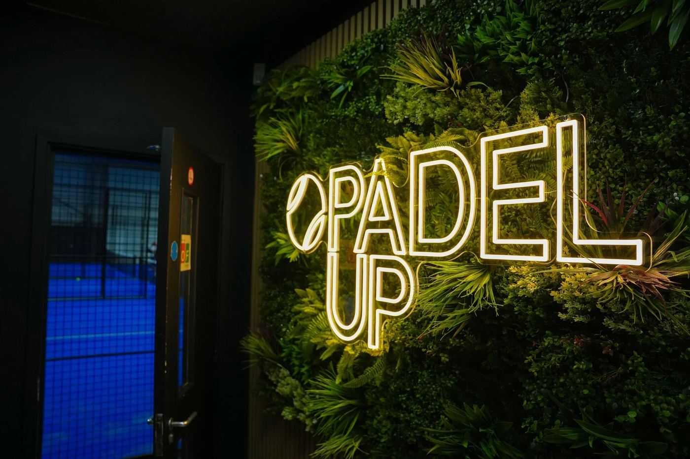 A neon Padel Up sign on a green plant wall beside a doorway onto a blue court
