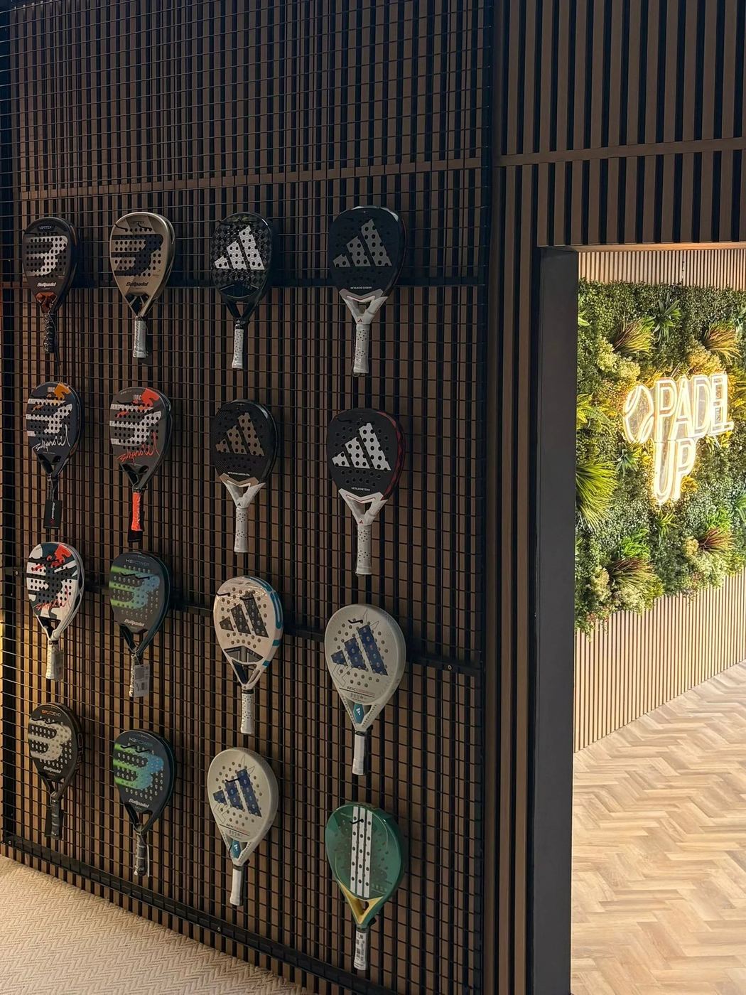 A wall of padel rackets displayed on dark slatted panels beside a leafy green sign