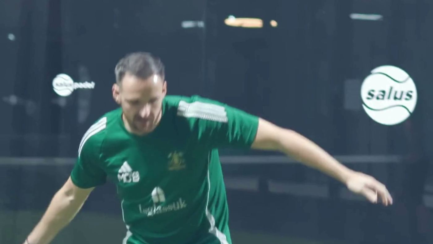 A player in a green shirt moving across the court during a game, with the Salus ball logo on the glass behind