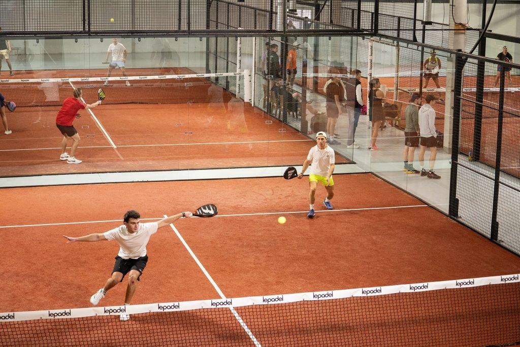 A doubles rally on an indoor padel court, with one player stretching low to reach the ball