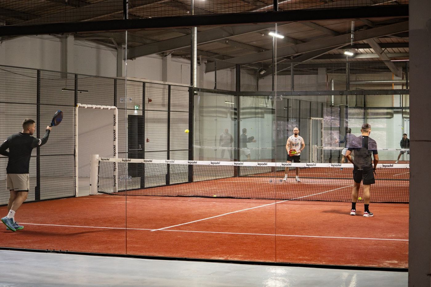 Players either side of the net on an indoor padel court with terracotta-coloured turf and glass walls