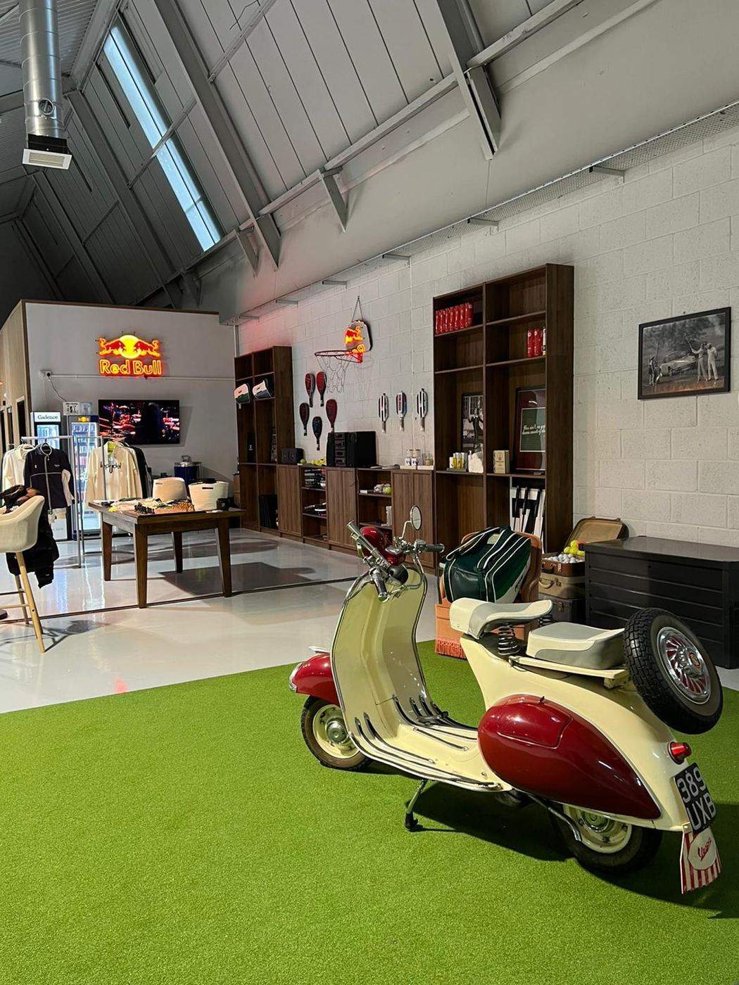 A lounge area inside the club with a vintage scooter on green artificial grass and shelves of padel gear behind