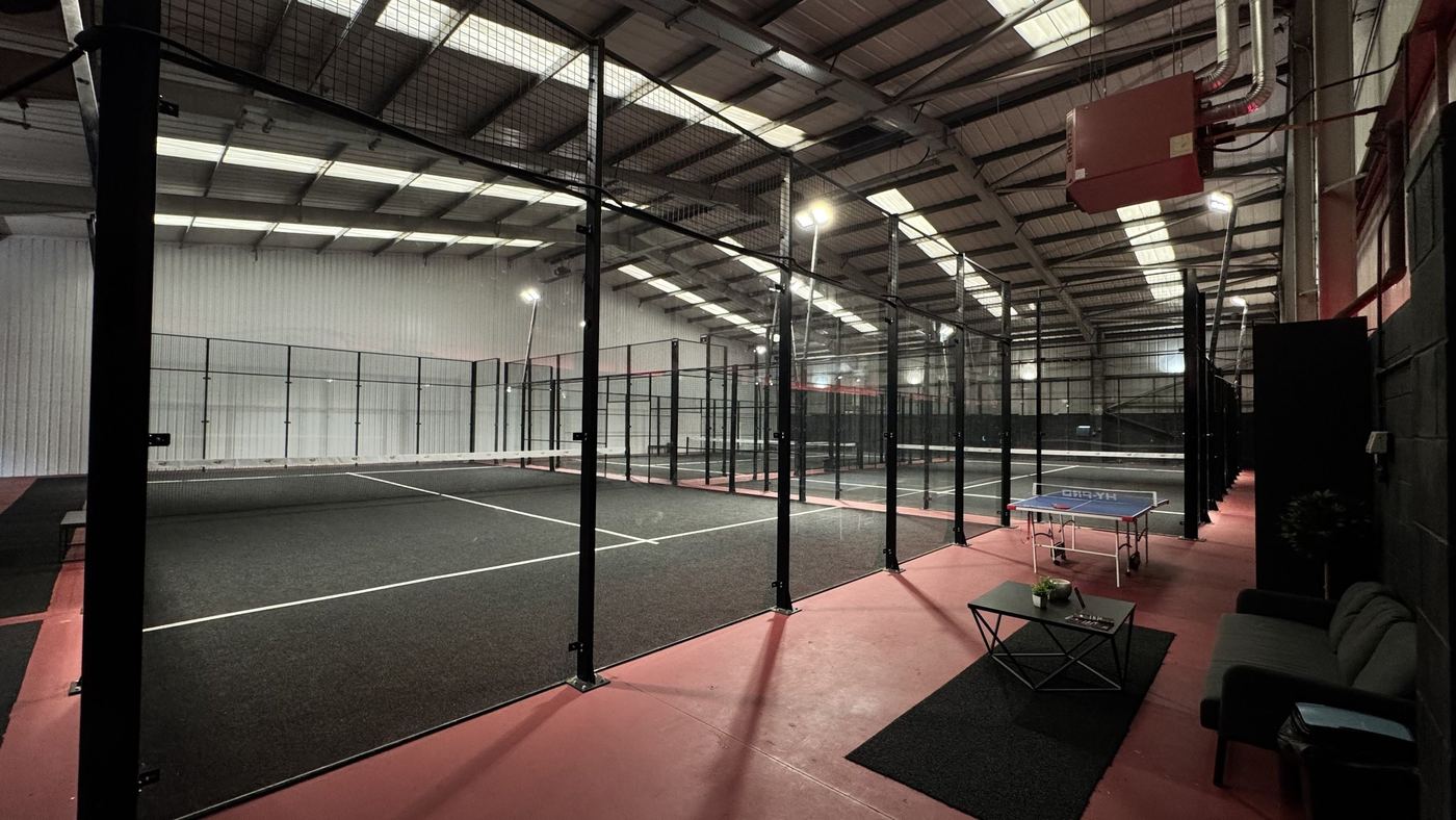 Padel courts behind glass and mesh in a large indoor hall with a courtside lounge and a table tennis table
