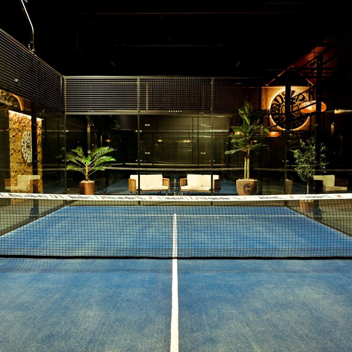 A blue padel court with lounge furniture visible through the glass at the far end