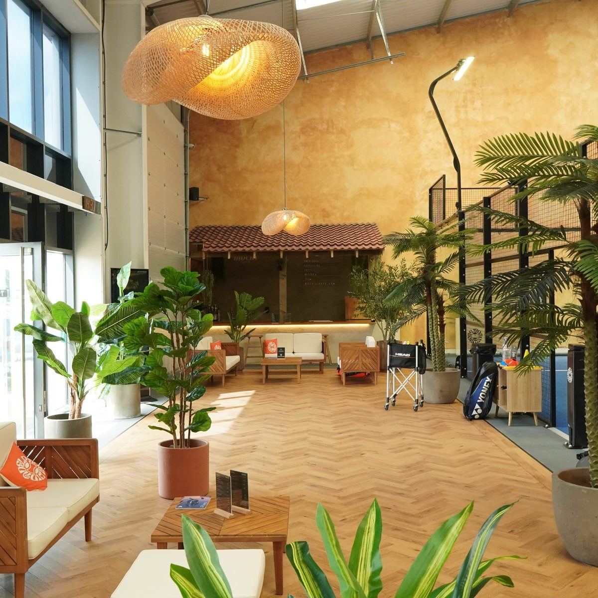 Cream sofas, wooden tables, plants and a woven pendant light in a club lounge beside the courts