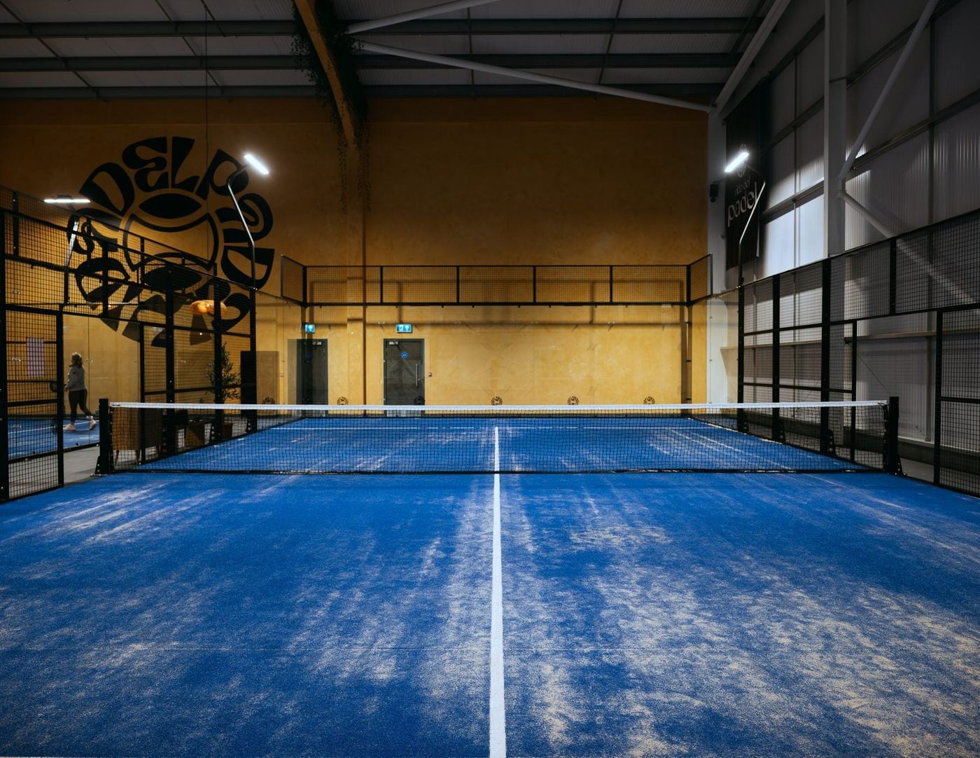 A blue padel court under a timber-beamed roof with an ochre wall carrying the club logo behind it